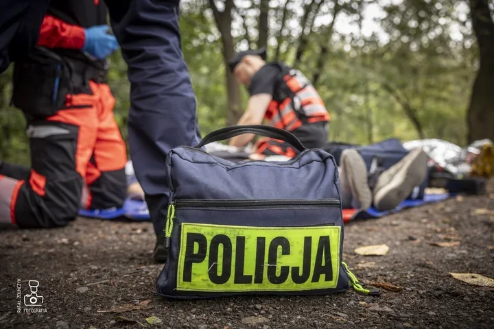 Funkcjonariusze policji udzielają pomocy poszkodowanemu leżącemu na ziemi w lesie, w pierwszym planie torba oznaczona napisem POLICJA.