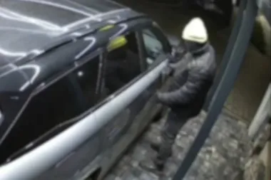 Mężczyzna w czarnej kurtce i jasnej czapce stoi przy czarnym SUV-ie i dotyka jego drzwi na brukowanej nawierzchni.